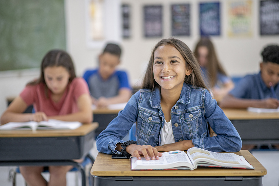 A beautiful female teenager is a high school student sitting at her desk in her classroom. She is wearing a denim jacket and is smiling widely.